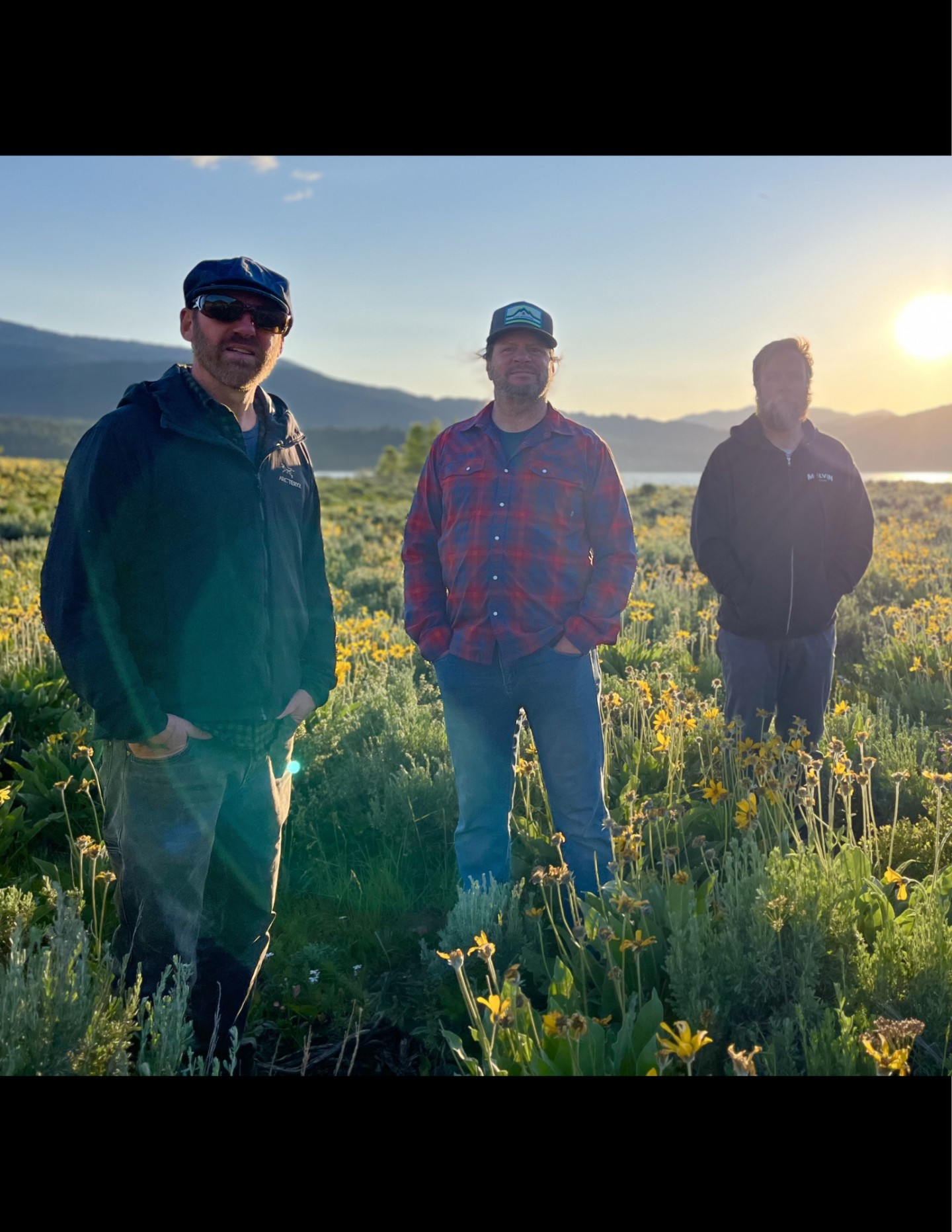 Three people in a sunlit field with mountains in the background.