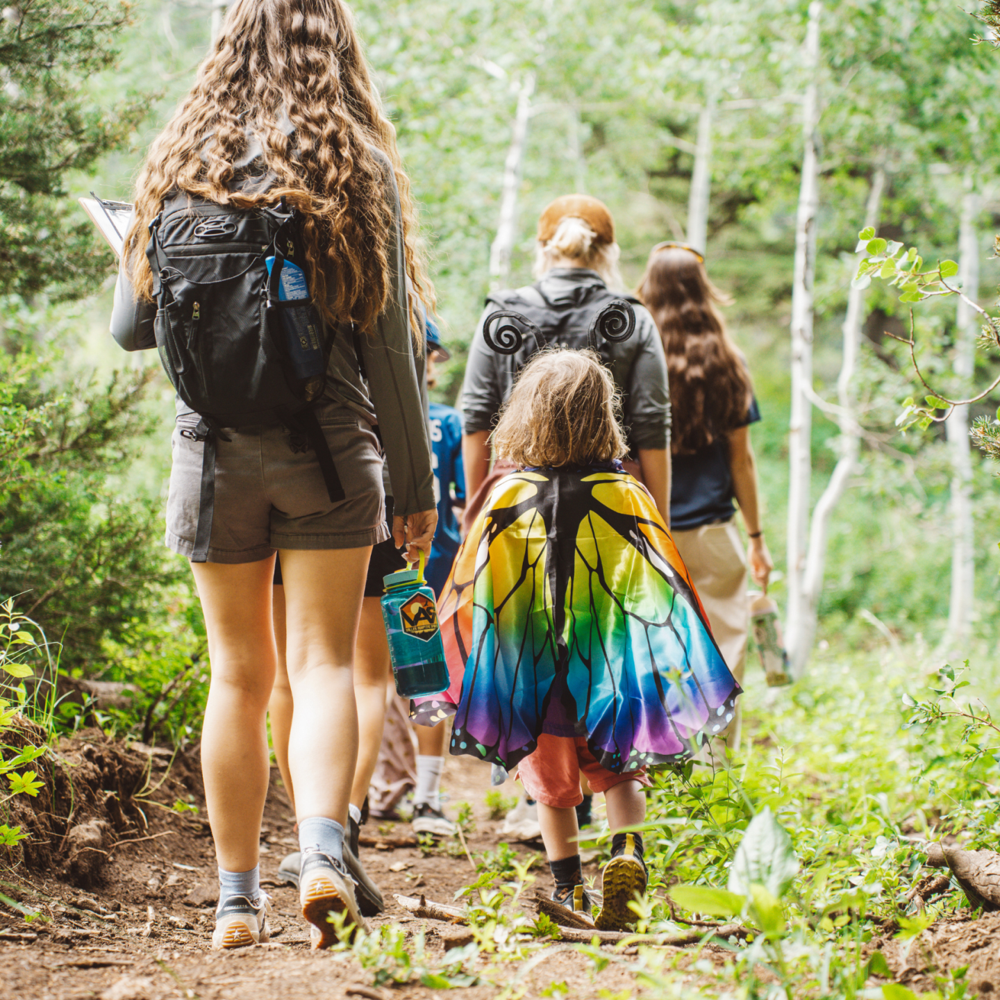 Group hiking on a forest trail; child wears colorful butterfly wings.