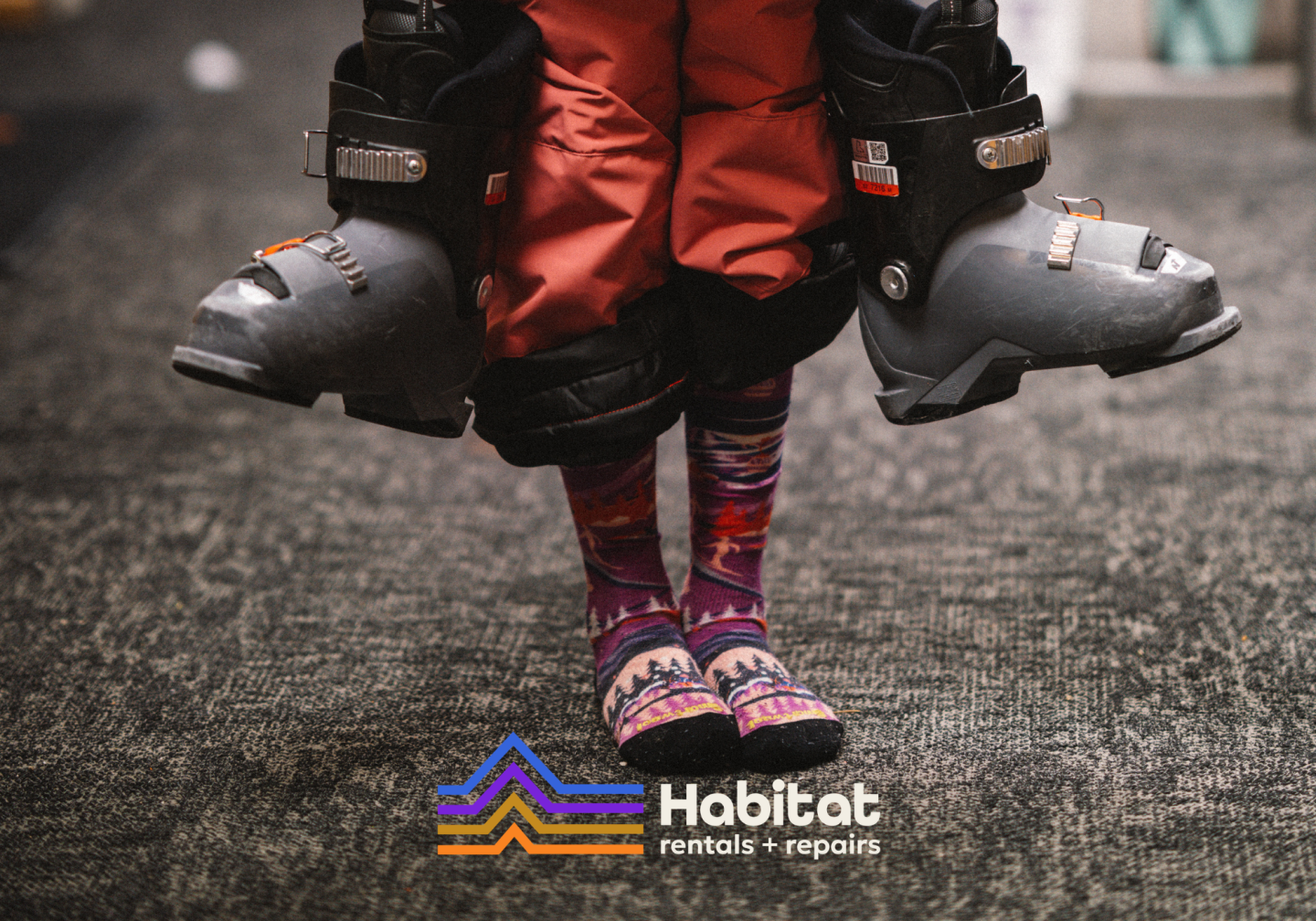 Child in red pants wearing ski boots, lifted above asphalt with colorful skis below.