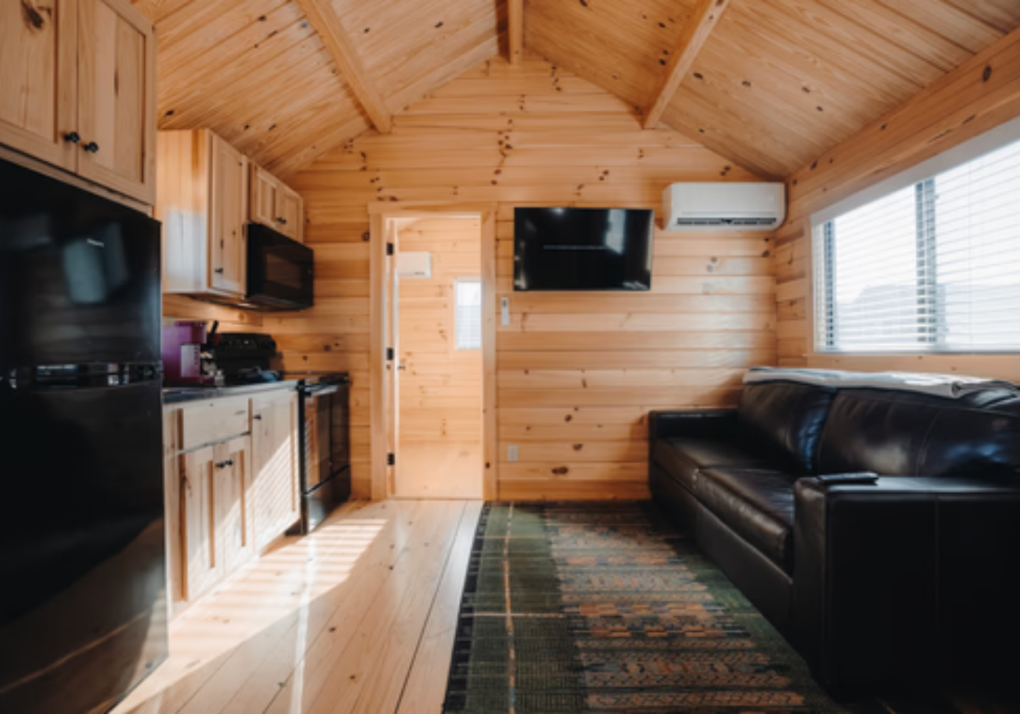 Cozy wood-paneled cabin interior with kitchen, black sofa, and wall-mounted TV.