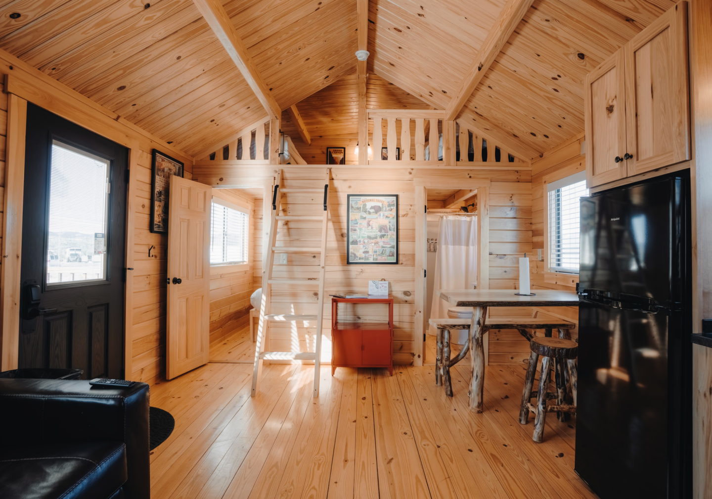 Cozy wood-paneled cabin interior with dining table, loft ladder, and black refrigerator.
