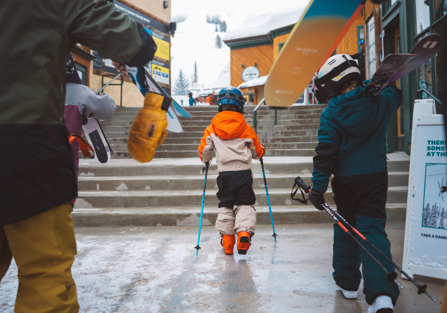 Child skiing downhill on a snowy village street, guided by adults.