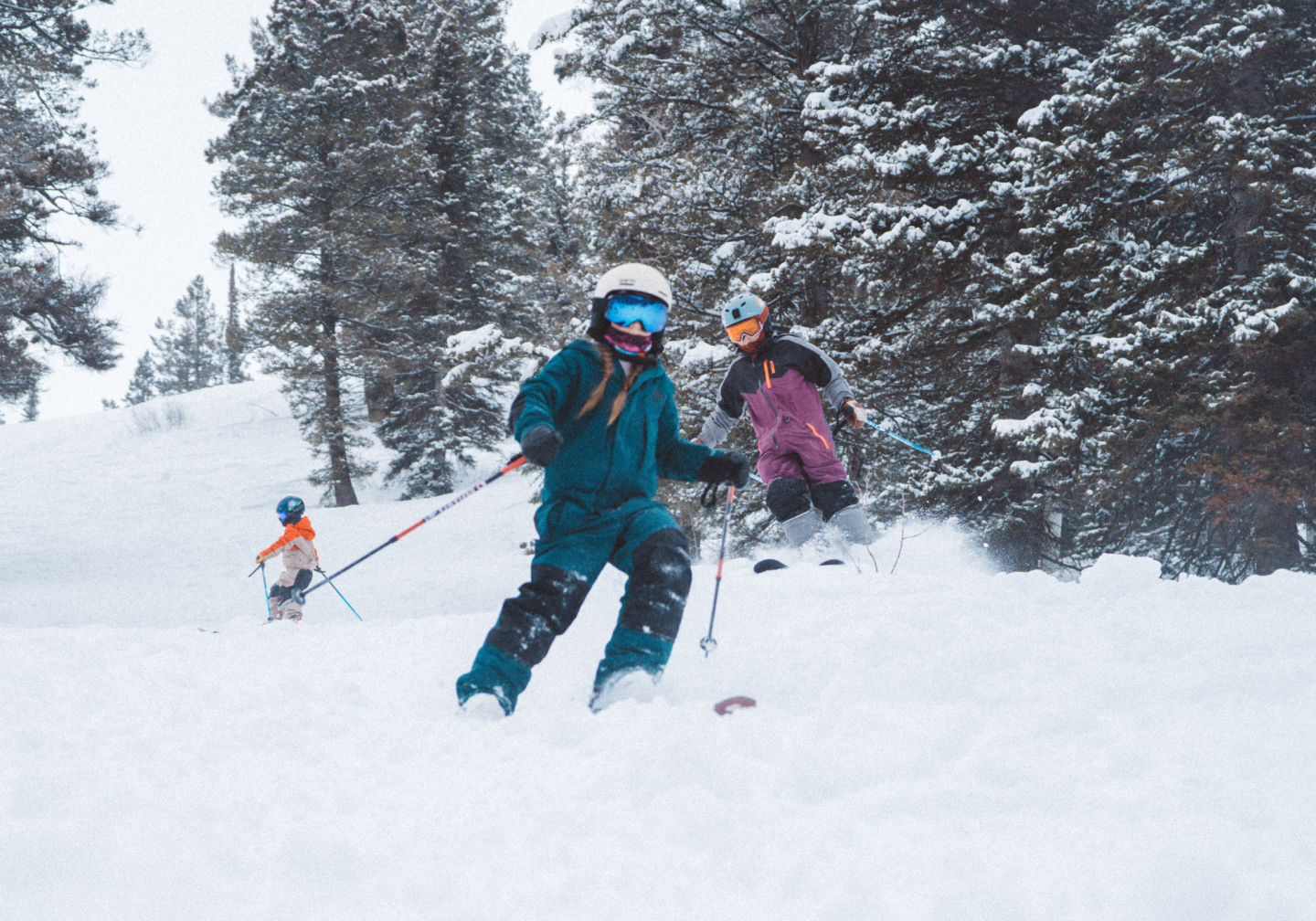 Two skiers in colorful jackets descend a snowy slope among pine trees.